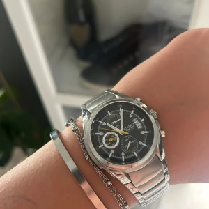 Casio kronografklocka i silver - Stilren Casio kronografklocka med svart urtavla, flera små ur och datumvisning. Klockan har en robust länk i silverfärgat metall och tydliga visare, inklusive en gul sekundvisare. Perfekt accessoar för dig som gillar klassisk och sportig stil.