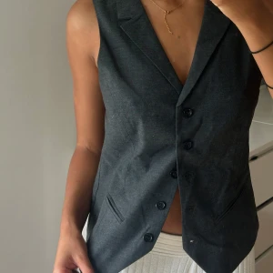 Mörkgrå kostymväst med knappar - Snygg mörkgrå kostymväst med klassisk krage och tre knappar framtill. Västen har två fuskfickor fram och justerbart spänne i ryggen. Framsidan är i matt tyg medan ryggen är i glansigare material. Perfekt för en stilren och trendig look.