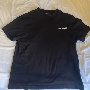 Svart t-shirt från Axel Arigato - Svart t-shirt från Axel Arigato med vit logga tryckt på bröstet. Klassisk rund halsringning och korta ärmar. T-shirten har en clean och stilren look, perfekt för dig som gillar minimalistisk design.