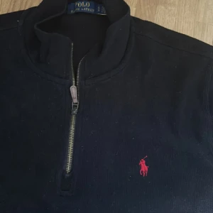 Svart half zip tröja Polo Ralph Lauren - Svart långärmad tröja från Polo Ralph Lauren med half zip och klassisk ståkrage. Tröjan har röd broderad logga på bröstet och är tillverkad i mjuk bomull. Perfekt för en clean och stilren look.