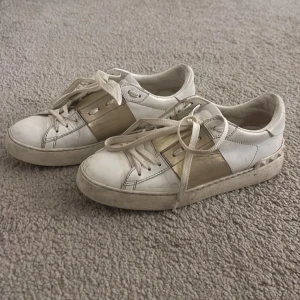 Valentino Open sneakers vit/guld strl 38 - Snygga Valentino Open sneakers i vitt skinn med bred guldfärgad detalj över mitten. Klassisk rund tå, platt sula och snörning framtill. Ikoniska nitar baktill på sulan ger extra edge. Perfekta för dig som gillar lyxiga och trendiga sneakers. Lite smutsiga men går att tvätta, inget som syns så mycket. 