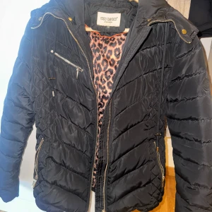 Svart pufferjacka med fuskpäls Roco Baroco - Svart pufferjacka från Roco Baroco med snyggt quiltat mönster och dragkedjor i silver. Jackan har en stor huva med beige fuskpäls och ett coolt leopardmönstrat innerfoder. Avtagbar päls. Perfekt för dig som vill ha en varm och trendig look i vinter.