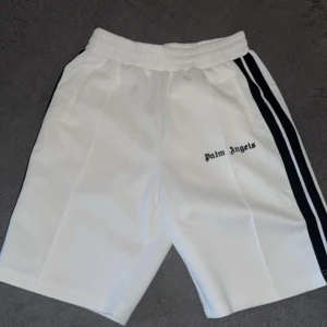 Palm Angels vita shorts med svarta ränder - Palm Angels vita shorts med svarta sidoremsor och broderad logga på framsidan. De har elastisk midja och är tillverkade i ett mjukt syntetmaterial som ger en clean och sportig vibe. Perfekta för dig som gillar streetwear och vill sticka ut.