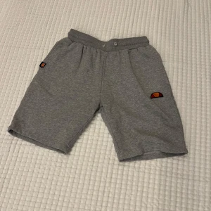 Grå Ellesse shorts med logga - Snygga grå shorts från Ellesse med stor vit logga och klassisk broderad emblem på ena benet. Mjuk bomullskvalitet, elastisk midja och avslappnad passform. Perfekta för chill eller sport, med skön känsla och stilren look.