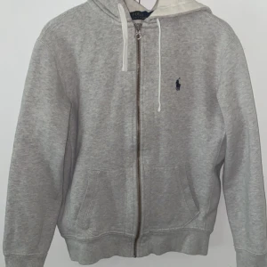 Grå hoodie från Polo Ralph Lauren - Snygg grå hoodie från Polo Ralph Lauren med dragkedja, huva med vita snören och klassisk blå logga på bröstet. Mjuk insida och två fickor framtill. Perfekt för chill dagar eller när du vill ha en clean och enkel stil.
