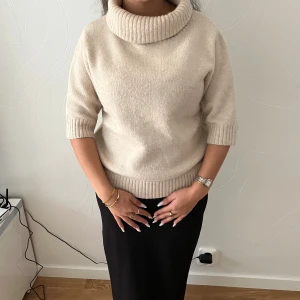 Beige polotröja från Indiska  - Säljer en beige polotröja med bred, ribbad krage och halvlånga ärmar. Tröjan är stickad i mjuk ullmix. Perfekt inför hösten och vintern. Är i storlek S men passar bra i storlek M
