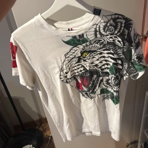 Vit t-shirt med tigertryck från Horspist - Säljer en vit t-shirt från Horspist med ett stort, detaljerat tigertryck i svart, grönt och rött framtill. Ena ärmen har en röd paljettpatch med logga. Skön passform och coola detaljer som verkligen sticker ut. Storlek S passar XS/S