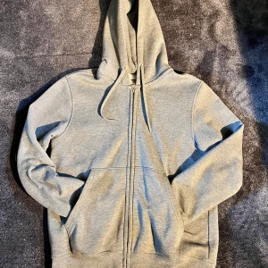 Grå hoodie med dragkedja och huva - En stilren grå hoodie med hel dragkedja framtill och huva med snörning. Tröjan har två stora fickor på magen och är långärmad. Perfekt för chill dagar eller när du vill ha en enkel och clean look.