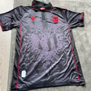 Svart Albanien fotbollströja Macron - Snygg svart fotbollströja från Albanien med röd FSHF-logga och röda detaljer. Tröjan har korta ärmar, krage med knapp och geometriskt mönster på framsidan. Tillverkad i lätt och ventilerande polyester, perfekt för match eller träning.