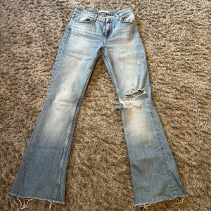 Ljusblå bootcut jeans med slitningar - Säljer ett par ljusblå bootcut jeans med slitna detaljer och hål vid knät. Jeansen har hög midja och råa kanter nertill för en avslappnad vibe. Perfekta för dig som gillar en trendig och lite edgy look.