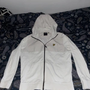 Vit hoodiejacka från Lyle & Scott - Vit hoodiejacka från Lyle & Scott med dragkedja och klassisk gul logga på bröstet. Jackan har huva, två fickor framtill och är tillverkad i mjukt bomullsmaterial. Perfekt för en chill och stilren look.