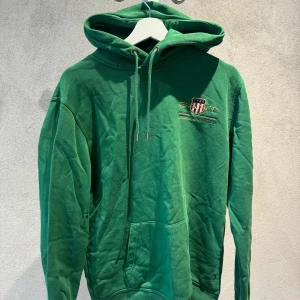 Grön hoodie från GANT med broderi - Säljer en grön hoodie från GANT i storlek L. Tröjan har huva med snörning, broderad logga och emblem på bröstet samt en stor magficka. Tillverkad i mjuk bomull, perfekt för chill dagar. Klassisk loose passform och långa ärmar.