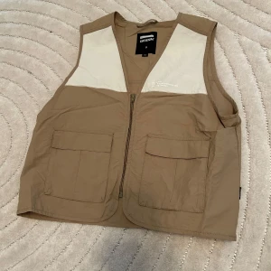Beige utility fiskeväst från Dr Denim - Säljer en beige utilityväst från Dr Denim med två stora fickor framtill och dragkedja. Västen har ljusa paneler upptill vid axlarna och är tillverkad i ett lätt polyestertyg. Perfekt för lager-på-lager och streetwear-vibe.