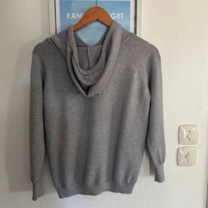Grå stickad hoodie med snörning - En stilren grå stickad hoodie med huva och snörning framtill. Tröjan är i xs. Perfekt för en chill och avslappnad look. Materialet känns mjukt och skönt mot huden. Hör av dig vid funderingar!