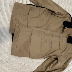 Beige parkajacka från Levi's - Snygg beige parkajacka från Levi's med svart krage i manchester. Jackan har stora praktiska fickor framtill och en mindre bröstficka. Perfekt för dig som gillar klassisk stil med coola detaljer och vill ha en jacka som funkar hela hösten. Har två fickor i bak som leder till ryggen, du kan ha ryggen som ryggsäck