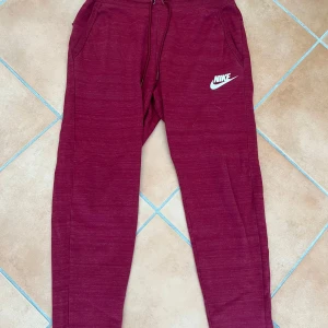 Vinröda mjukisbyxor från Nike - Snygga vinröda mjukisbyxor från Nike med vit logga på låret. Byxorna har dragsko i midjan och två sidofickor. Perfekta för chill eller träning, med en skön och avslappnad passform.