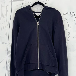 Mörkblå hoodie med dragkedja från H&M - Mörkblå hoodie från H&M i storlek M med hel dragkedja framtill och två stora fickor. Tröjan har huva och långa ärmar, tillverkad i mjuk bomull som är skön att ha på sig. Perfekt för chill dagar eller när du vill ha en enkel och stilren look.