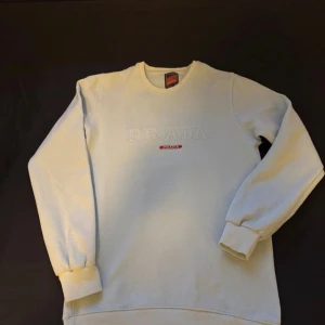 Vit sweatshirt från Prada - Säljer en clean vit sweatshirt från Prada med broderad logga framtill och diskret röd detalj. Tröjan har rund hals, långa ärmar och breda muddar vid ärmslut och nederkant. Perfekt för dig som gillar stilrena designer och exklusiva märken.