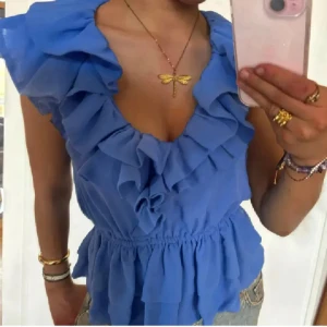 Blå volangblus från H&M - Supersöt blå blus från H&M med volanger längs v-ringningen och axlarna. Blusen är ärmlös och har resår i midjan för en snygg passform. Tillverkad i ett lätt och luftigt polyestertyg, perfekt för varma dagar när du vill känna dig extra fin. Storlek 40 men är lite mindre i storleken! 