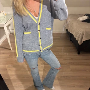 Grå kofta med gula detaljer - Säljer en grå kofta med gula ränder längs kanterna och på ärmarna. Koftan har v-ringning, knappar framtill och två fickor med gul kant. Stickad i mjukt material och har långärmade ärmar. Perfekt för dig som gillar färg och chill stil.