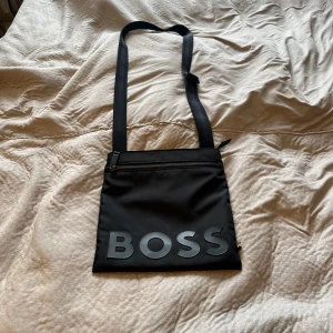 Svart axelväska från BOSS - Snygg svart axelväska från BOSS med justerbar rem och stort BOSS-tryck framtill. Väskan har en dragkedja upptill och är tillverkad i slitstark polyester. Perfekt för att bära det viktigaste när du är på språng.