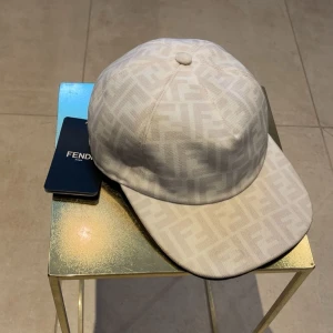 Vit Fendi keps  - Snygg vit keps från Fendi med det klassiska FF-monogrammet i ett diskret, ton-i-ton mönster över hela kepsen. Tillverkad i 100% polyester och levereras med original ask och dustbag. Perfekt för dig som vill ha en lyxig och stilren keps. Pris kan diskuteras vid snabb affär. Hör av er vid minsta funderingar!✅📦