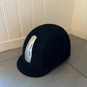 Svart ridhjälm CRW med silverdetalj - Snygg svart ridhjälm från CRW med en cool silverfärgad detalj framtill. Hjälmen har en klassisk rundad form och är tillverkad i ett mjukt, sammetliknande material. Perfekt för dig som gillar ridning och vill ha en stilren look i stallet. Ny & aldrig använd.