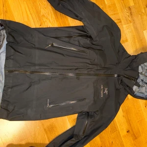 Svart vindjacka från Arc'teryx S - Svart vindjacka från Arc'teryx i storlek S. Jackan har huva, flera dragkedjeförsedda fickor och en clean look. Perfekt för dig som gillar outdoor och vill ha något lätt och smidigt. Materialet är syntet och jackan har långa ärmar. Gore-tex