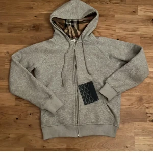 Grå Burberry hoodie med dragkedja - Snygg grå hoodie från Burberry med klassiskt rutigt foder i huvan och broderad logga på bröstet. Tröjan har dragkedja, snörning i huvan och ribbade muddar. Perfekt för dig som vill ha en stilren och lyxig vardagslook. Använd 1 gång