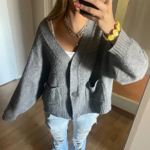 Grå oversized stickad kofta - Mysig grå stickad kofta med oversized passform och stora knappar framtill. Koftan har två fickor och bred ribbad kant nedtill och vid ärmsluten. Perfekt att slänga över en t-shirt för en chill look.