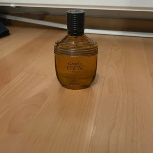 Gary's Den Le Parfum Fomo parfym - Parfymflaska i bärnstensfärgat glas med svart ribbad kork. Flaskan har en rundad form och texten 'Gary's Den Le Parfum' på framsidan. Perfekt för dig som gillar unika dofter och snygg design på hyllan. Dior homme intense liknande och nästan bättre. 