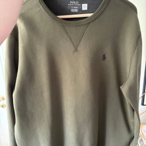 Olivgrön sweatshirt från Polo Ralph Lauren - Snygg olivgrön sweatshirt från Polo Ralph Lauren med klassisk rund hals och blå broderad logga på bröstet. Tröjan har långa ärmar och är gjord i mjuk bomull. Enkel och stilren design som funkar till det mesta. Nypris är 1700, den är använd 3-4 gånger. Storlek är Large plussize, den passar för XL. Säljer för 700 men priset är förhandlingsbart.