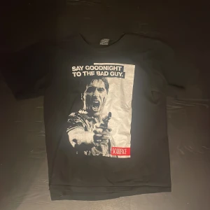 Svart Scarface t-shirt med tryck - Svart t-shirt med stort Scarface-tryck framtill och texten 'Say goodnight to the bad guy'. T-shirten har korta ärmar och klassisk rund hals. Materialet är mjuk bomull och passformen är normal. Perfekt för dig som gillar film och streetwear.