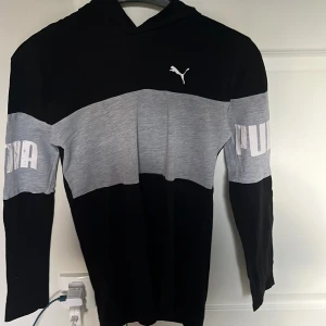 Svart och grå hoodie från Puma XL - Snygg hoodie från Puma i svart och grått med vit logga på bröstet och stora PUMA-tryck på ärmarna. Tröjan har huva, lång ärm och är tillverkad i mjukt material som passar perfekt till chill eller träning.