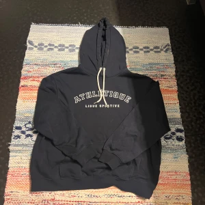 Mörkblå hoodie från H&M med tryck - Säljer en mörkblå hoodie från H&M med vit ATHLETIQUE-tryck på bröstet och snörning i huvan. Tröjan har långa ärmar, ribbade muddar och en klassisk känguruficka framtill. Perfekt för chill dagar eller när du vill ha en sportig vibe.