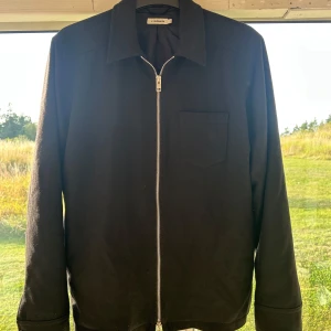 overshirt  - Stilren svart overshirt från J.Lindeberg med rak passform och klassisk krage. Jackan har en bröstficka och hel dragkedja framtill. Perfekt att slänga på över en hoodie eller t-shirt för en clean look. Skulle säga att passformen är mellan s-m 