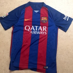 Barcelona tröja 2017 - Säljer en officiell FC Barcelona matchtröja från Nike med breda blå och röda ränder, klubbmärke på bröstet och Qatar Airways-tryck. Kortärmad modell med rund hals och La Liga-märke på ärmen. Perfekt för dig som älskar fotboll och Barca.