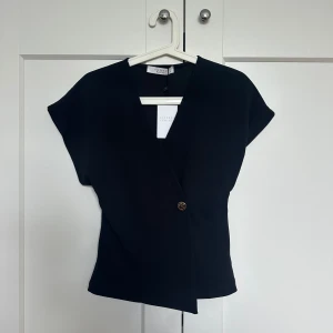 Svart omlott topp - En super fin svart omlott topp i tjockare siden material! En super fin guld knapp på framsidan som går att knäppa upp! Oanvänd med lappen på!! 