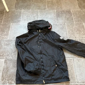 Svart vindjacka från Moncler med huva - Svart vindjacka från Moncler med snygg huva som har röda och vita ränder. Jackan har dragkedja framtill, snörning i huvan och Moncler-logga på ärmen. Tillverkad i glansig polyester och har praktiska fickor med tryckknappar. Perfekt för dig som vill ha en clean och sportig look.