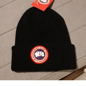 Svart ribbad mössa Canada Goose - Svart ribbstickad mössa från Canada Goose med klassisk rund logotyp framtill. Perfekt för kalla dagar och snygg till alla outfits. Tjockt uppvik för extra värme och stil.