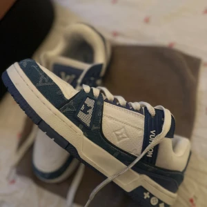 Blåvita sneakers i jeanstyg  - Säljer ett par sneakers i blått och vitt med klassiska loggor och detaljer i jeanstyg precis som originalet. Skorna har snörning, rund tå och platt sula. Ovandelen är i en mix av skinn och textil med monogramdetaljer. Perfekta för dig som vill sticka ut med lyxig streetstyle. Använda ca 2 gånger så väldigt fint skick! 