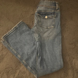 Blå bootcut jeans med fickdetalj  - Snygga blå jeans med bootcut passform och klassisk femficksdesign. Jeansen har en cool ficka med knapp bak och ljus tvätt som ger en avslappnad vibe. Perfekta för dig som gillar en lite vidare siluett nertill. Materialet är klassisk denim.