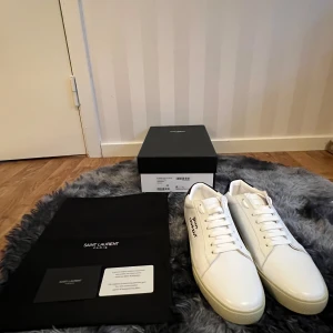 Saint Laurent skor - Exklusiva vita sneakers från Saint Laurent i slät skinn med svart detalj på hälen och handskriven logga på sidan. Klassisk låg modell med rund tå, platt sula och vita snören. Tillverkade i Italien och levereras med originalkartong och dustbag.