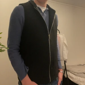 Svart väst från ASOS med dragkedja - Svart väst från ASOS i enkel och stilren design. Västen har en rak passform, hög krage och hel dragkedja framtill. Tillverkad i ett mjukt material med slät insida. Perfekt att slänga över en hoodie eller tröja för en clean look.