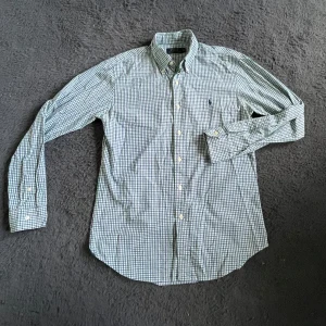 Ralph Lauren skjorta  - Snygg rutig skjorta från Polo Ralph Lauren i grönt, blått och vitt. Klassisk button-down krage, långa ärmar och knappar framtill. Perfekt för dig som gillar en tidlös och fräsch stil. Skjortan är i mjuk bomull och har en broderad logga på bröstet.