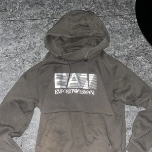 Mörkgrå EA7 Emporio Armani hoodie - Snygg mörkgrå hoodie från EA7 Emporio Armani med stor logga i silver på bröstet. Tröjan har huva med snörning och en klassisk känguruficka framtill. Perfekt för dig som gillar streetwear och vill ha en stilren look.