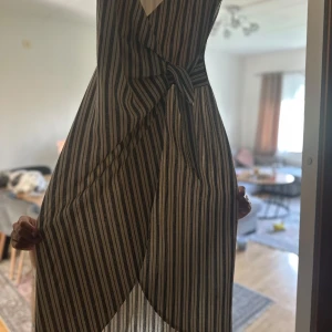 Randig omlottklänning med knyt - Snygg omlottklänning med svart och beigea ränder. Klänningen har knyt i midjan och ett luftigt fall, perfekt för varma dagar. Den är ärmlös och har en stilren, somrig look som passar dig som gillar trendiga detaljer.