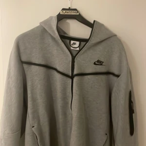 Grå Nike hoodie med dragkedja - Snygg grå hoodie från Nike med svart logga på bröstet och svarta detaljer längs dragkedjan och ärmarna. Tröjan har huva, hel dragkedja och praktisk ficka med dragkedja på ena ärmen. Perfekt för chill eller träning. Kan även passa M