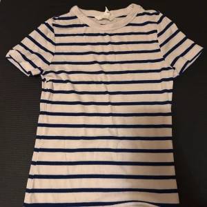 Vit och blå randig t-shirt från H&M - Vit t-shirt med blå horisontella ränder från H&M. Modellen har rund hals och korta ärmar. Materialet är mjuk ribbad bomull som känns skön mot huden. Perfekt för dig som gillar klassisk och enkel stil med en twist.