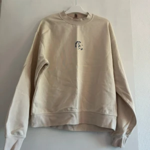 Beige sweatshirt från H&M XS - Enkel och stilren beige sweatshirt från H&M i storlek XS. Tröjan har rund halsringning, lång ärm och en liten svart broderad detalj på bröstet. Perfekt för en chill och avslappnad look. Tillverkad i mjuk bomullsmix.